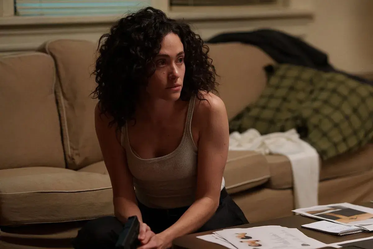 Kobieta z kręconymi włosami, w podkoszulku, siedzi na podłodze, trzymając pistolet. Scena z filmów i seriali Emmy Rossum.