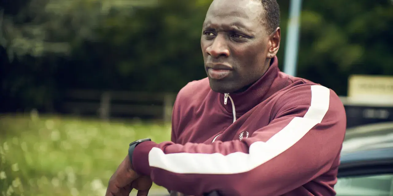 Omar Sy w bordowej bluzie z białymi pasami, gotowy do akcji w kolejnych filmach, serialach i programach.
