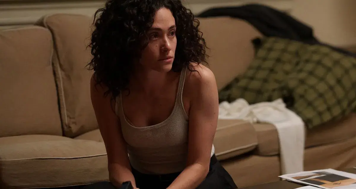 Kobieta z kręconymi włosami, w podkoszulku, siedzi na podłodze, trzymając pistolet. Scena z filmów i seriali Emmy Rossum.
