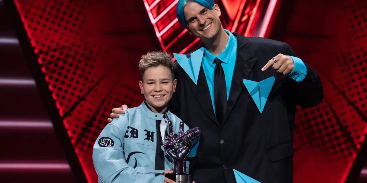 Chłopiec z błękitną kurtką i trofeum w kształcie dłoni, obok mężczyzny z niebieskimi włosami. To on wygrał The Voice Kids!