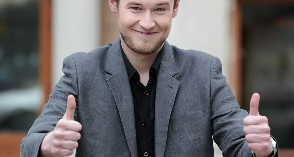 Krzysztof Wójcik z "Milionerów" pokazuje kciuki w górę, uśmiechając się.