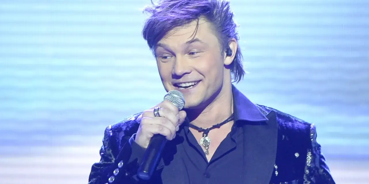 Jaka to melodia piosenkarze: przystojny mężczyzna z fioletowymi włosami śpiewa do mikrofonu w błyszczącej marynarce.