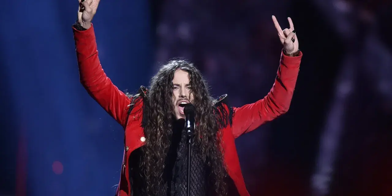 Michał Szpak na Eurowizji w czerwonej marynarce, z uniesionymi rękami i gestem rock'n'rolla.