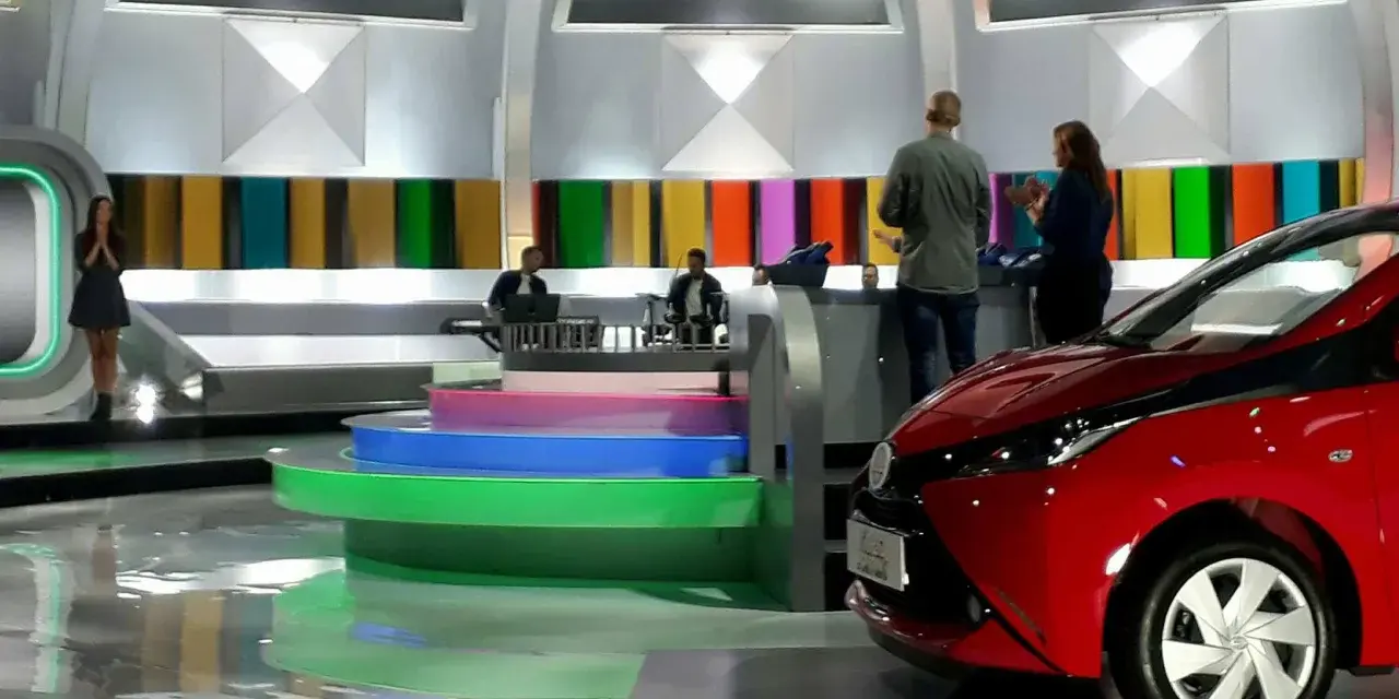Studio telewizyjne, gdzie nagrywane jest koło fortuny. Widać scenografię z kolorowymi panelami, schodami i czerwonym samochodem.