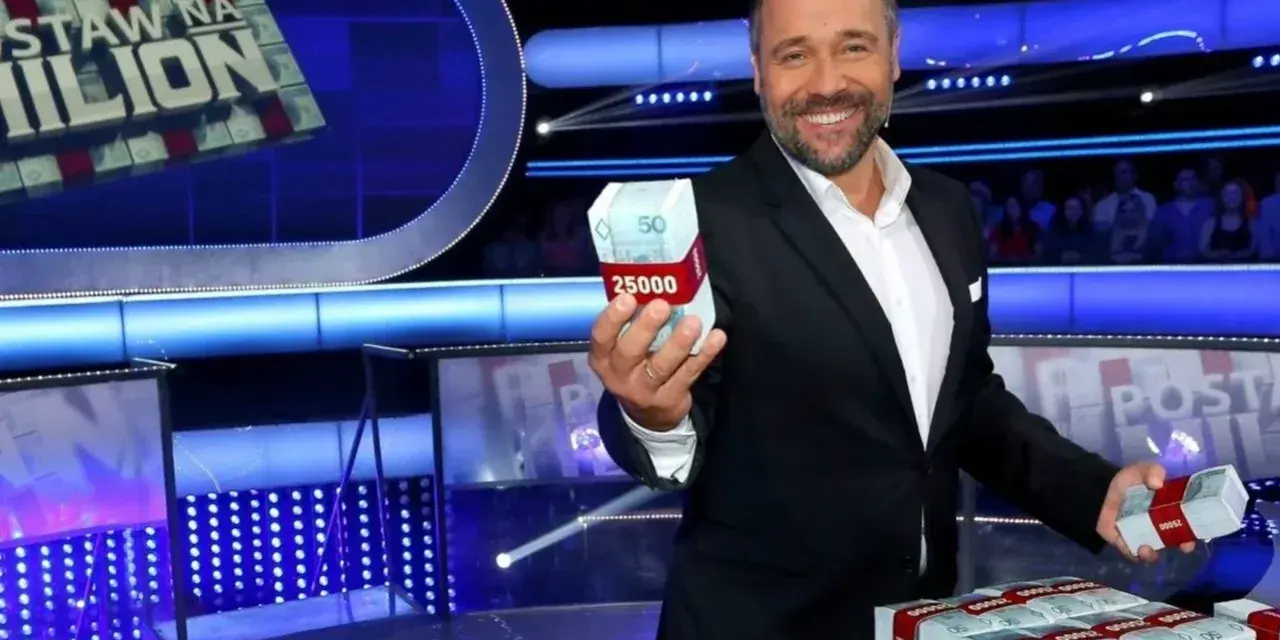Uśmiechnięty prowadzący teleturniej trzyma w dłoni paczkę pieniędzy. Na stole piętrzą się stosy banknotów.