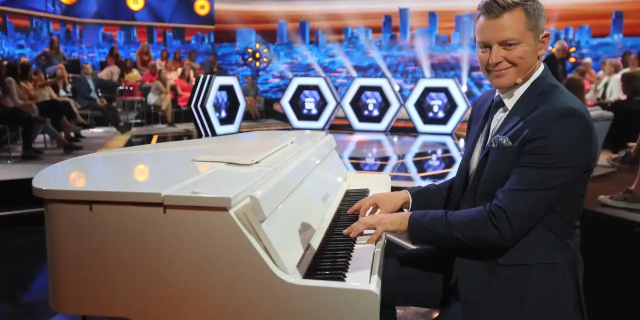 Mężczyzna gra na białym pianinie w studio telewizyjnym. Czy to melodia z programu "Jaka to melodia"?