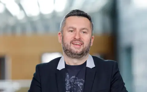 Uśmiechnięty mężczyzna w czarnej marynarce i t-shircie z nadrukiem. Ciekawe, ile zarabia Norbi w Kole Fortuny?