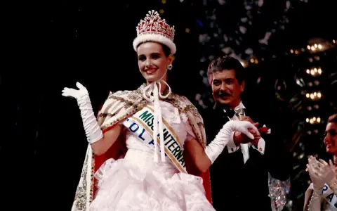 Uśmiechnięta Miss International w koronie i białej sukni, z szarfą "Miss Polonia". Obok niej mężczyzna w smokingu.