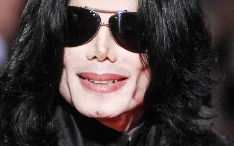 Michael Jackson w okularach przeciwsłonecznych i czarnych włosach. Jego uśmiech i twarz budzą pytania, jak zmarł Michael Jackson.