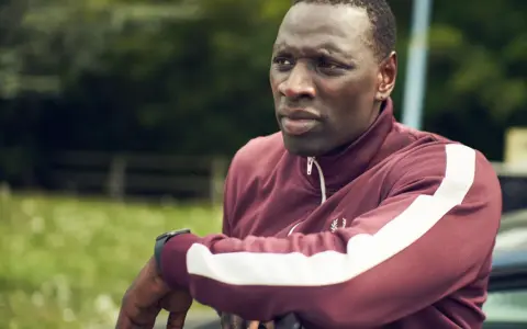 Omar Sy w bordowej bluzie z białymi pasami, gotowy do akcji w kolejnych filmach, serialach i programach.