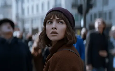 Młoda kobieta w brązowym płaszczu i bordowej czapce patrzy w górę, otoczona tłumem. Czy to zapowiedź netflix nowości dzisiaj?