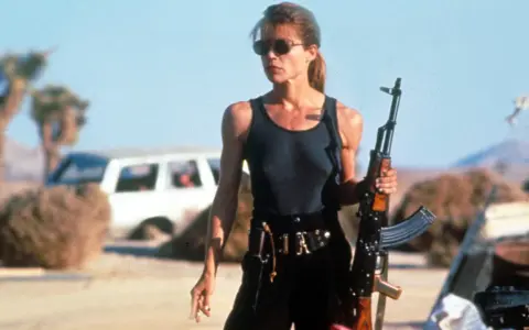 Linda Hamilton w roli Sary Connor, gotowa do walki w pustynnym krajobrazie. Jej ikoniczne role w filmach i serialach na zawsze zapisały się w historii kina.