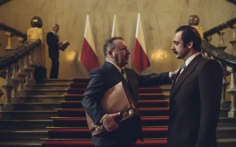 Hubert Niedziela, w garniturze, trzyma teczkę, rozmawiając z mężczyzną na schodach przed polskimi flagami.