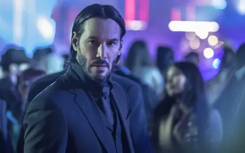 Keanu Reeves w garniturze, w tle rozmyte światła i tłum. Ikona kina akcji, znana z filmów takich jak John Wick.