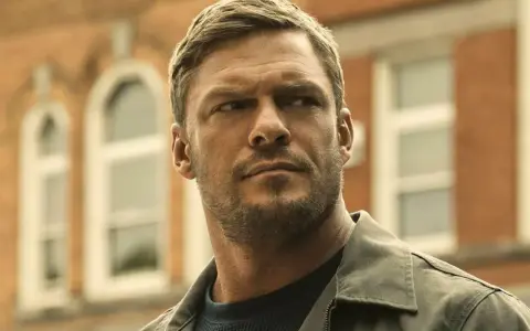 Alan Ritchson jako Jack Reacher. W tle ceglany budynek z białymi oknami.