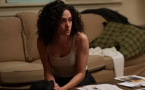 Kobieta z kręconymi włosami, w podkoszulku, siedzi na podłodze, trzymając pistolet. Scena z filmów i seriali Emmy Rossum.