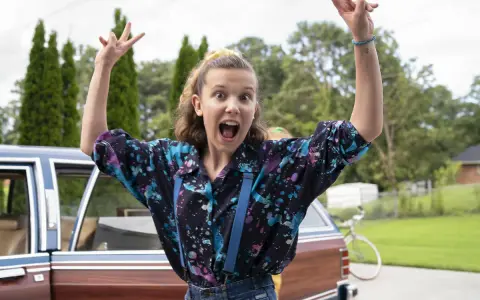 Millie Bobby Brown w kolorowej koszuli i szelkach, radośnie pozuje przy samochodzie. Jej filmy, seriale i programy to gwarancja emocji!