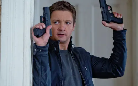 Jeremy Renner w akcji, jakiego znamy z jego filmów i seriali. W rękach trzyma dwa pistolety, gotowy do strzału.