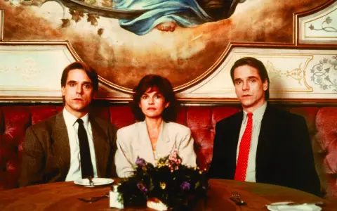 Jeremy Irons w filmach, tutaj z dwiema kobietami, siedzi przy stole w eleganckiej restauracji.