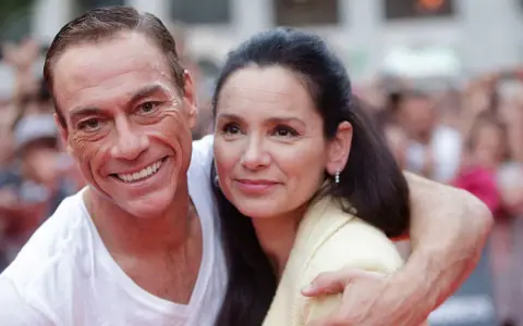 Jean-Claude Van Damme z uśmiechem obejmuje swoją żonę, Gladys Portugues, na tle tłumu.