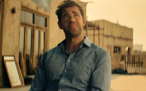 John Krasinski w niebieskiej koszuli, zamyślony. Widzimy fragmenty jego filmów, seriali i programów.