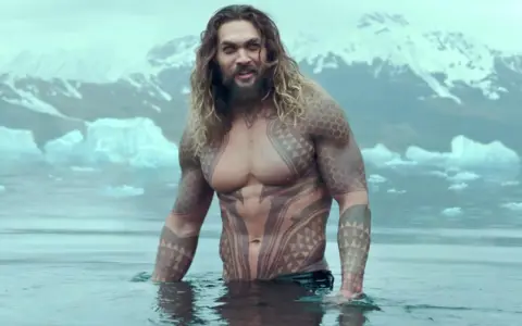 Jason Momoa w wodzie, z tatuażami, jak w filmach i serialach. W tle góry i lód.