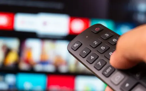 Dłoń trzyma pilot do telewizora, wybierając film na ekranie. To idealny moment na relaks z web tv.