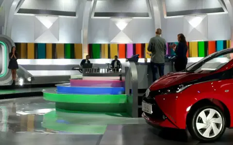 Studio telewizyjne, gdzie nagrywane jest koło fortuny. Widać scenografię z kolorowymi panelami, schodami i czerwonym samochodem.