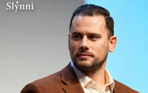Grzegorz Collins, znany z programów telewizyjnych, w eleganckim brązowym garniturze. Jego wiek jest trudny do określenia, ale prezentuje się dojrzale.