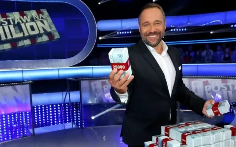 Uśmiechnięty prowadzący teleturniej trzyma w dłoni paczkę pieniędzy. Na stole piętrzą się stosy banknotów.