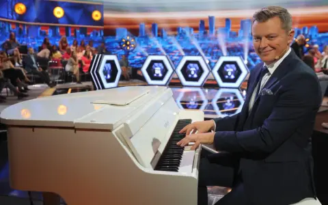 Mężczyzna gra na białym pianinie w studio telewizyjnym. Czy to melodia z programu "Jaka to melodia"?