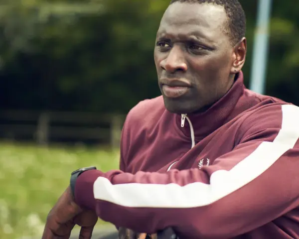 Omar Sy w bordowej bluzie z białymi pasami, gotowy do akcji w kolejnych filmach, serialach i programach.
