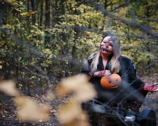 Kobieta w makijażu jack-o'-lantern trzyma dynię, gotowa na noc pełną **horrorów Netflix** w jesiennym lesie.