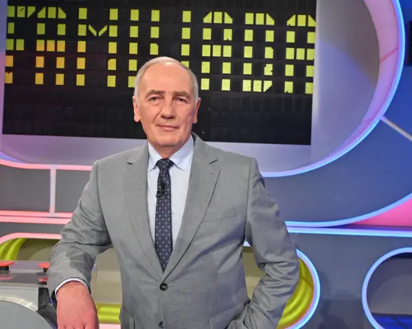 Mężczyzna w garniturze na tle planszy teleturniejowej, wyglądający na siłach i gotowy do gry, by być zdrowym jak Familiada.