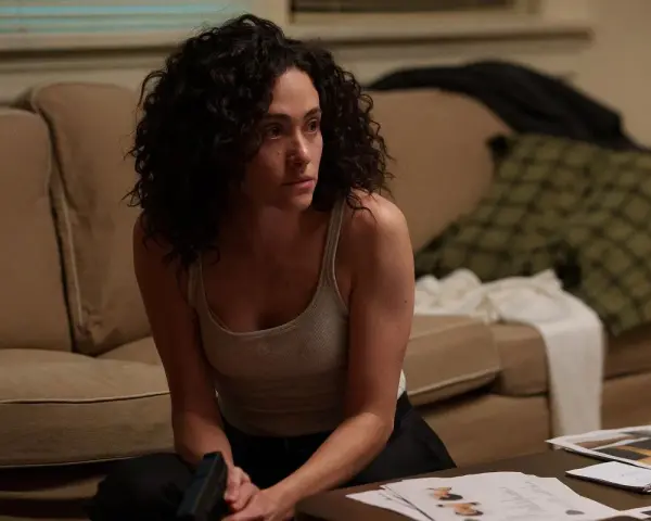 Kobieta z kręconymi włosami, w podkoszulku, siedzi na podłodze, trzymając pistolet. Scena z filmów i seriali Emmy Rossum.