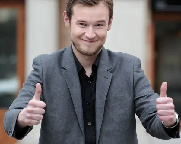 Krzysztof Wójcik z "Milionerów" pokazuje kciuki w górę, uśmiechając się.