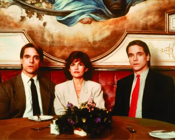 Jeremy Irons w filmach, tutaj z dwiema kobietami, siedzi przy stole w eleganckiej restauracji.