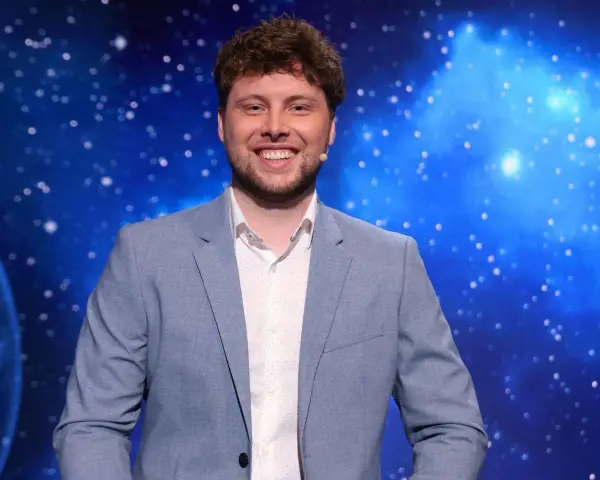 Uśmiechnięty mężczyzna w jasnej marynarce, jeden z dziesięciu jak się zgłosić, na tle kosmicznego nieba.