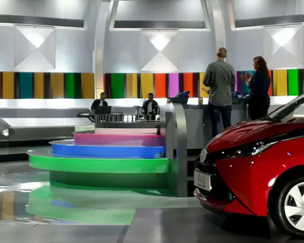 Studio telewizyjne, gdzie nagrywane jest koło fortuny. Widać scenografię z kolorowymi panelami, schodami i czerwonym samochodem.