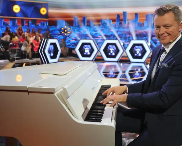Mężczyzna gra na białym pianinie w studio telewizyjnym. Czy to melodia z programu "Jaka to melodia"?