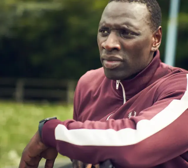 Omar Sy w bordowej bluzie z białymi pasami, gotowy do akcji w kolejnych filmach, serialach i programach.
