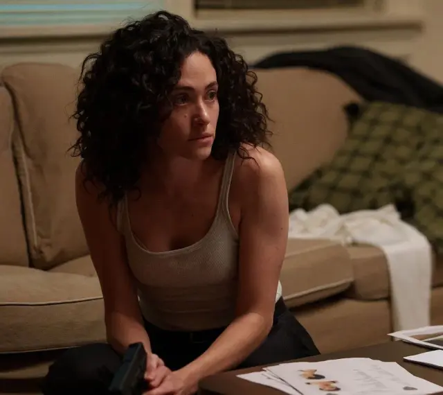 Kobieta z kręconymi włosami, w podkoszulku, siedzi na podłodze, trzymając pistolet. Scena z filmów i seriali Emmy Rossum.
