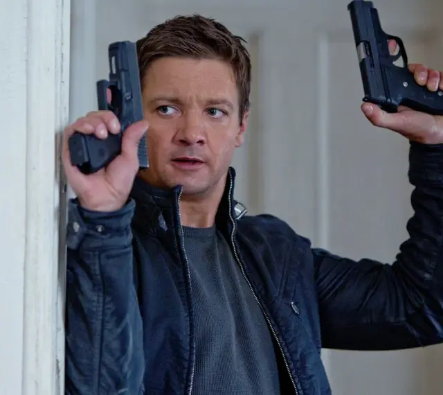 Jeremy Renner w akcji, jakiego znamy z jego filmów i seriali. W rękach trzyma dwa pistolety, gotowy do strzału.