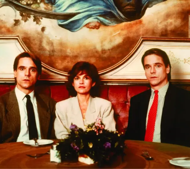 Jeremy Irons w filmach, tutaj z dwiema kobietami, siedzi przy stole w eleganckiej restauracji.