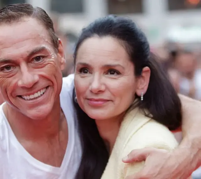 Jean-Claude Van Damme z uśmiechem obejmuje swoją żonę, Gladys Portugues, na tle tłumu.
