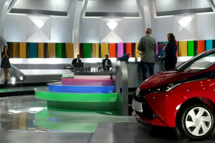 Studio telewizyjne, gdzie nagrywane jest koło fortuny. Widać scenografię z kolorowymi panelami, schodami i czerwonym samochodem.