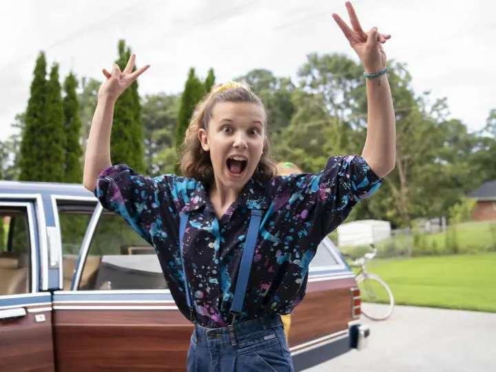 Millie Bobby Brown w kolorowej koszuli i szelkach, radośnie pozuje przy samochodzie. Jej filmy, seriale i programy to gwarancja emocji!