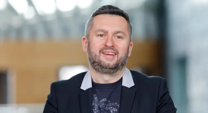 Uśmiechnięty mężczyzna w czarnej marynarce i t-shircie z nadrukiem. Ciekawe, ile zarabia Norbi w Kole Fortuny?