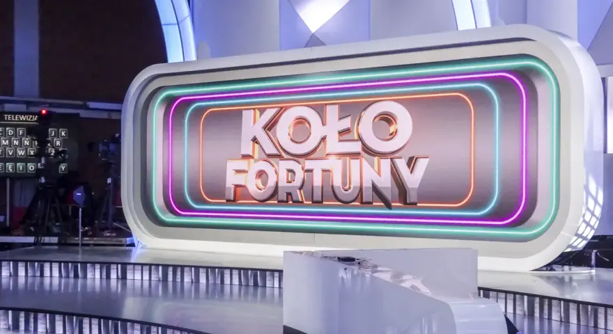 Na scenie studia telewizyjnego widnieje napis "KOŁO FORTUNY" otoczony neonami. To miejsce, gdzie marzenia o wygranej stają się rzeczywistością.