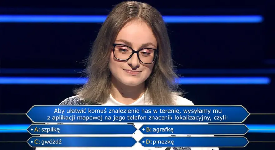 Młoda kobieta w okularach na tle niebieskich pasów, zastanawia się nad pytaniem z teleturnieju: "Aby ułatwić komuś znalezienie nas w terenie, wysyłamy mu z aplikacji mapowej na jego telefon znacznik lokalizacyjny, czyli: B: agrafkę".